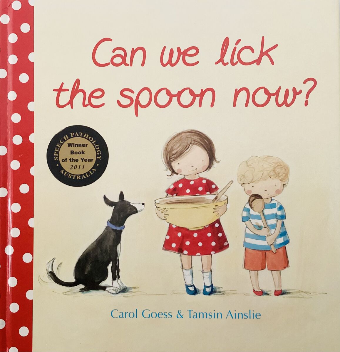 Cover of Can We Lick the Spoon Now? by Carol Goess and Tamsin Ainslie — a girl in a red polka dot dress holds a mixing bowl while a boy licks a wooden spoon and a black dog watches, with the 2011 Speech Pathology Australia Book of the Year award sticker